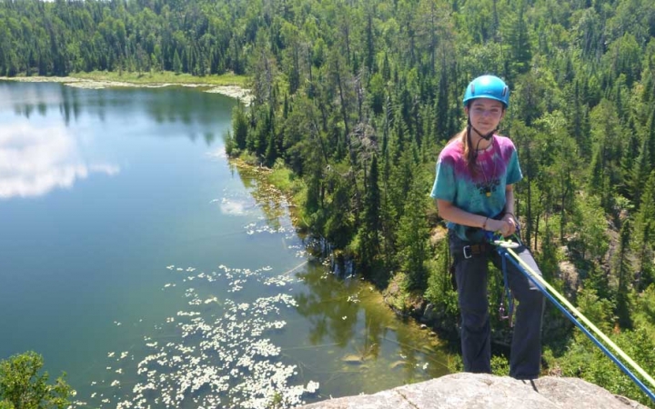 A person wearing safety gear is secured by ropes as they look at the camera and smile while rock climbing. They are high above a body of water and a green forrest. 